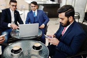 grupo de homem de negócios indiano de terno sentado no escritório no café com laptop, mensagens de texto em telefones e fazendo foto de café.