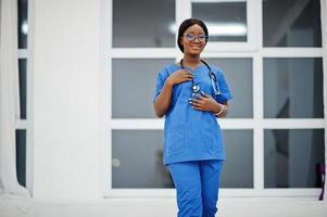 retrato de feliz feminino americano africano jovem médico pediatra no casaco uniforme azul e estetoscópio contra a janela no hospital. saúde, médico, especialista em medicina - conceito.