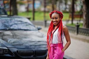 garota afro-americana na moda em calças cor de rosa e dreads vermelhos posou ao ar livre contra o carro preto. foto