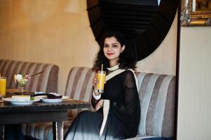 jovem indiana usar no elegante saree preto sentado no restaurante com um copo de suco na mão. foto
