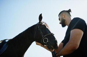 homem árabe de barba alta usa preto com cavalo árabe. foto