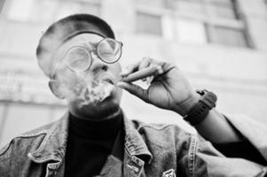 feche o retrato do homem afro-americano na jaqueta jeans, boina e óculos, fumando charuto. foto