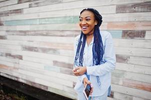 menina afro-americana elegante com dreads segurando o celular na mão, ao ar livre contra a parede de madeira. foto