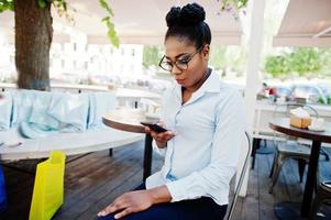 garota afro-americana usa óculos com celular sentado no caffe ao ar livre. foto