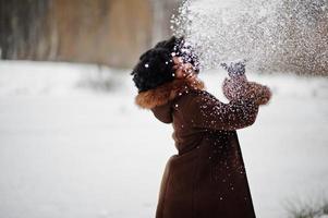 mulher afro-americana de cabelo encaracolado usa casaco de pele de carneiro e luvas posadas no dia de inverno vomita neve. foto