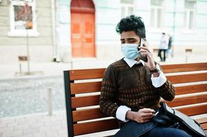conceito de coronavírus covid-19. homem indiano do sul da Ásia usando máscara para proteger do vírus corona sentado no banco com telefone celular. foto