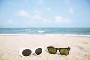 dois óculos de sol da moda fecham na areia da praia no verão. bela vista para o mar papel de parede de céu azul claro, férias de verão relaxe o fundo com espaço de cópia. óculos de sol viajam conceito de amantes de casal. foto