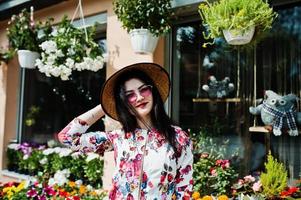 retrato de verão da menina morena de óculos cor de rosa e chapéu contra loja de flores. foto