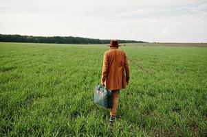 homem elegante de óculos, jaqueta marrom e chapéu com bolsa posou em campo verde. foto