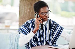 homem africano em roupas tradicionais e óculos sentado atrás do laptop no caffe ao ar livre e falando no celular. foto