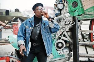 homem afro-americano na jaqueta jeans, boina e óculos, fumando charuto e posou contra o antigo veículo retrô. foto