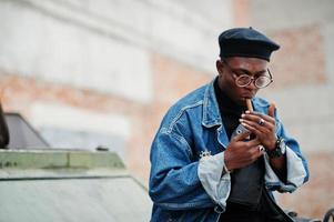 homem afro-americano de jaqueta jeans, boina e óculos, acende um charuto e posou contra o veículo blindado militar btr. foto