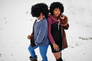 duas mulheres afro-americanas de cabelo encaracolado usam casaco de pele de carneiro e luvas posadas no dia de inverno, falando ao telefone. foto