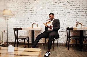 forte e poderoso homem afro-americano de terno preto sentado no café, ler o livro e cheirar a flor no tubo de ensaio. foto