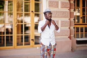 homem afro-americano elegante na camisa branca e calça colorida com chapéu e óculos posou ao ar livre. menino modelo preto elegante. foto