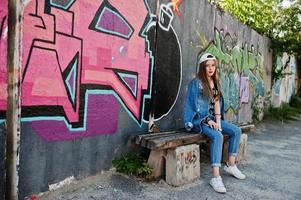 menina elegante casual hipster em boné e jeans usa música de fones de ouvido do telefone celular contra grande parede de graffiti com bomba. foto