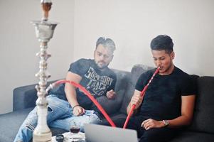 dois homens indianos bonitos e elegantes relaxando no quarto e fumando narguilé. foto