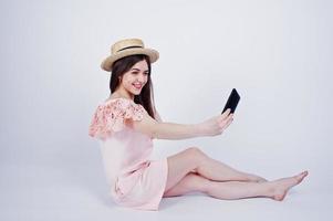 retrato de uma mulher elegante de vestido rosa e chapéu sentado no chão e tomando selfie no estúdio. foto