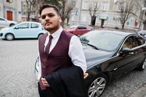 elegante empresário indiano com roupa formal em pé contra carro preto na rua da cidade. foto