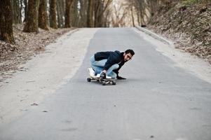 homem árabe de estilo de rua em óculos com longboard longboard na estrada. foto