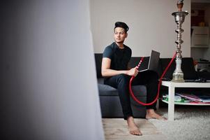 homem indiano bonito e elegante em preto sentado no quarto, fumando narguilé e trabalhando no laptop. foto