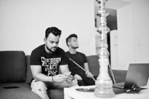 dois homens indianos bonitos e elegantes relaxando no quarto e fumando narguilé. um deles olhando para o celular. foto