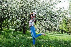 retrato de primavera de menina morena de óculos cor de rosa e chapéu no jardim de flor verde. foto