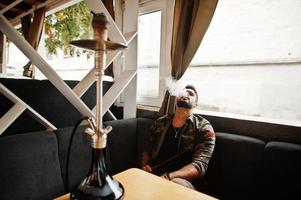 homem árabe de barba elegante de óculos e jaqueta militar fumando cachimbo de água no bar da rua. modelo árabe descansando. foto