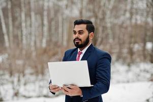homem de negócios de barba indiana elegante de terno posou em dia de inverno ao ar livre com o laptop nas mãos. foto