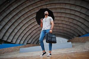 homem de barba árabe alto na moda usar camisa, jeans e óculos de sol com guarda-chuva e bolsa posada no salão de arena. foto