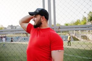 jovem homem musculoso barbudo brutal usar camisa vermelha, shorts e boné no estádio. foto