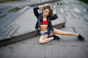 garota sexy modelo encaracolado no top vermelho, shorts jeans, jaqueta de couro e tênis posou no chão. foto