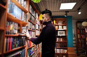 homem alto e inteligente estudante árabe, use gola rulê violeta e óculos, na biblioteca selecionando livro nas prateleiras. foto