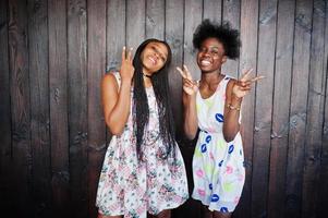 duas namoradas africanas negras em vestidos de verão posaram contra um fundo escuro de madeira e mostrando dois dedos. foto