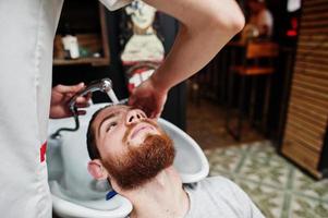 jovem barbudo lavando a cabeça pelo cabeleireiro enquanto está sentado na cadeira na barbearia. alma de barbeiro. foto