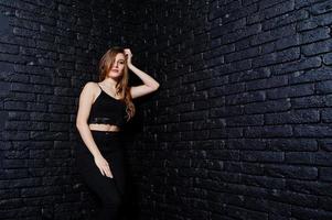 menina morena bonita usa preto, posando no estúdio contra a parede de tijolos escuros. retrato de modelo de estúdio. foto