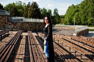 retrato de menina morena de óculos cor de rosa usar preto posou ao ar livre em dia ensolarado contra a linha de bancos. foto