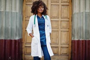 médico americano africano fêmea no jaleco com estetoscópio ao ar livre contra a porta da clínica.