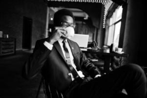 homem afro-americano de negócios usa terno preto e óculos no escritório, bebendo café da manhã antes do dia de trabalho. foto