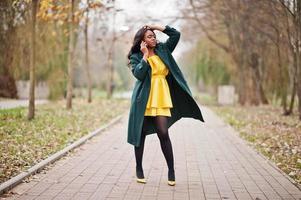 mulher afro-americana elegante no casaco verde e vestido amarelo posou contra o parque outono e falando no telefone. foto