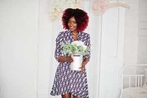 elegante modelo americano africano alto com cabelo afro vermelho vestido posado no quarto branco com vaso de flores nas mãos. foto