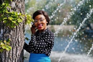 menina elegante modelo americano africano de óculos, saia azul e blusa preta posou fontes de fundo ao ar livre. foto