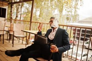 bonito homem árabe bem vestido fumar charuto com copo de uísque na varanda do pub. foto