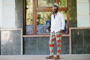 homem afro-americano elegante na camisa branca e calça colorida com chapéu e óculos posou ao ar livre. menino modelo preto elegante. foto