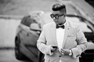 homem árabe elegante de jaqueta, gravata borboleta e óculos escuros contra carro suv preto. empresário rico árabe com telefone celular. foto