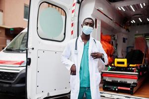 paramédico masculino africano em máscara médica protetora facial em frente ao carro da ambulância. foto