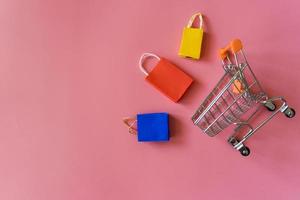 conceito on-line de compras mínimo, saco de compras de papel colorido e carrinho descem do fundo rosa flutuante para espaço de cópia. cliente pode comprar tudo de casa e o mensageiro vai entregar. foto