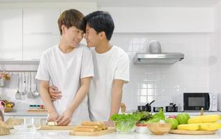 afetuoso jovem casal masculino asiático tendo um tempo romântico juntos durante o cozimento da salada de pão na cozinha branca com um sorriso no rosto. conceito de vida doméstica lgbt. foco seletivo. foto