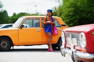bela dama afro-americana com sacos de compras em pé perto de carro retrô clássico laranja. foto
