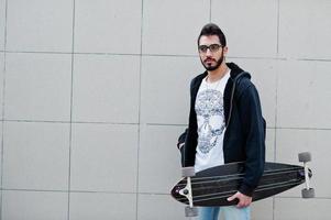 homem árabe de estilo de rua em óculos com longboard posou contra a parede cinza. foto